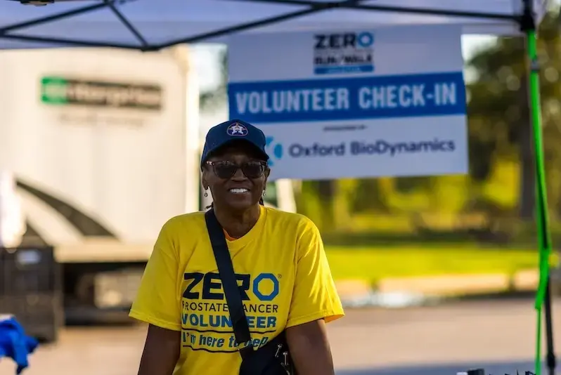 A Black female volunteer smiles at the race check-in area, wearing a yellow ZERO Prostate Cancer volunteer shirt. A Black female volunteer smiles at the race check-in area, wearing a yellow ZERO Prostate Cancer volunteer shirt.
