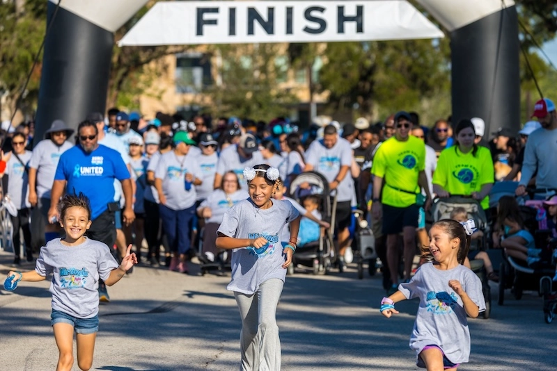 Race participants making their way towards the camera at the start of the race with three smiling girls running in the lead. Race participants making their way towards the camera at the start of the race with three smiling girls running in the lead.