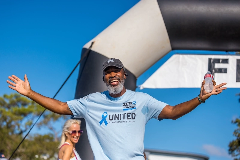 A Black male race participant holds his arms out in celebration at the finish line, water bottle in hand. A Black male race participant holds his arms out in celebration at the finish line, water bottle in hand.